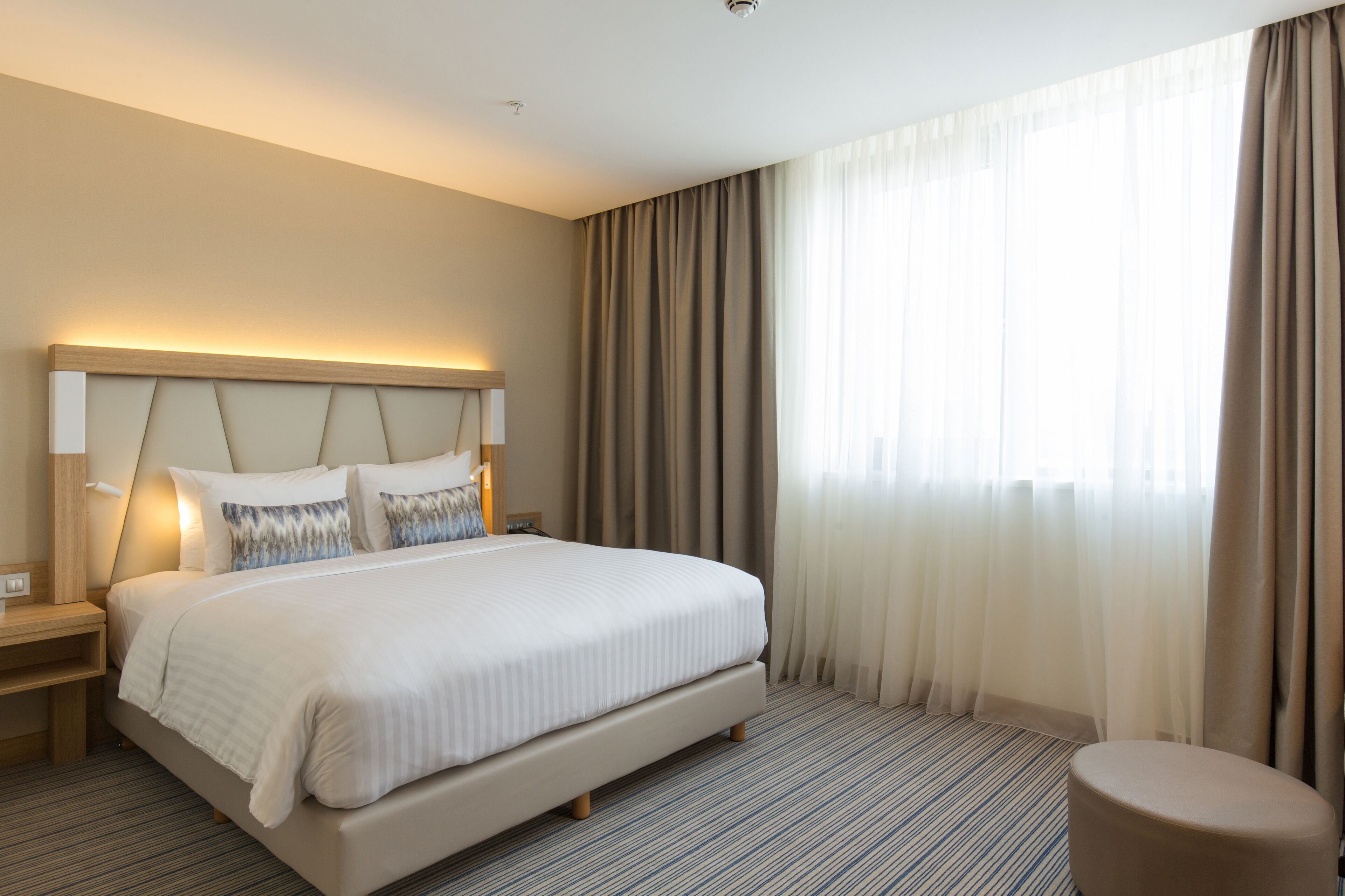 Ein Hotelzimmer mit einem großen Bett, einem Nachttisch, einem runden Hocker und einem großen Fenster mit Vorhängen.