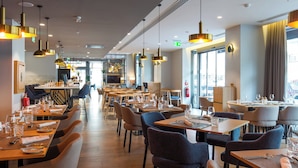 Ein modernes Restaurant mit mehreren Tischen, Stühlen, großen Fenstern und Pendelleuchten.