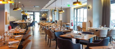 A modern restaurant with multiple dining tables and chairs, large windows, and pendant lighting.