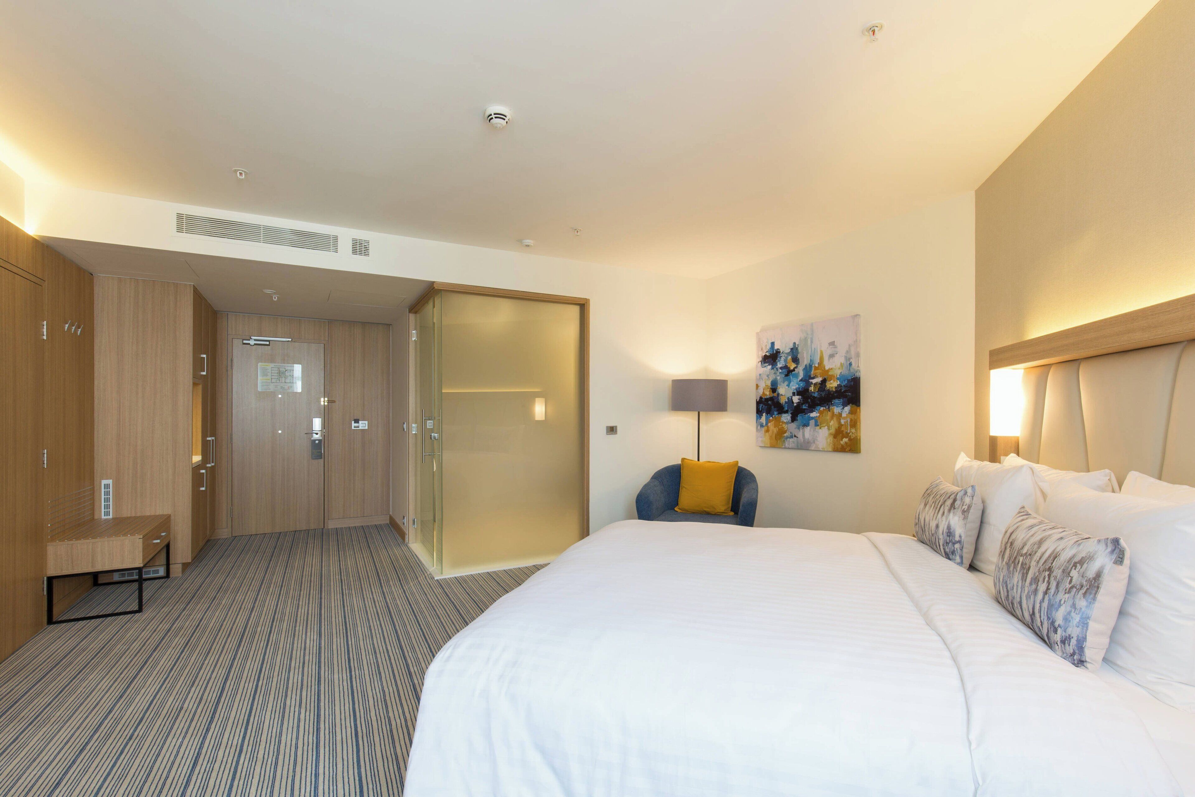 A hotel room with a large bed, a desk, and a chair. There is a painting on the wall and a lamp on the desk.