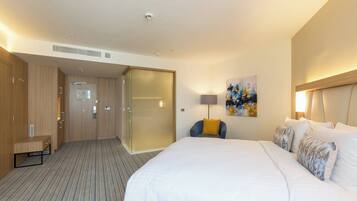 A hotel room with a large bed, a desk, and a chair. There is a painting on the wall and a lamp on the desk.