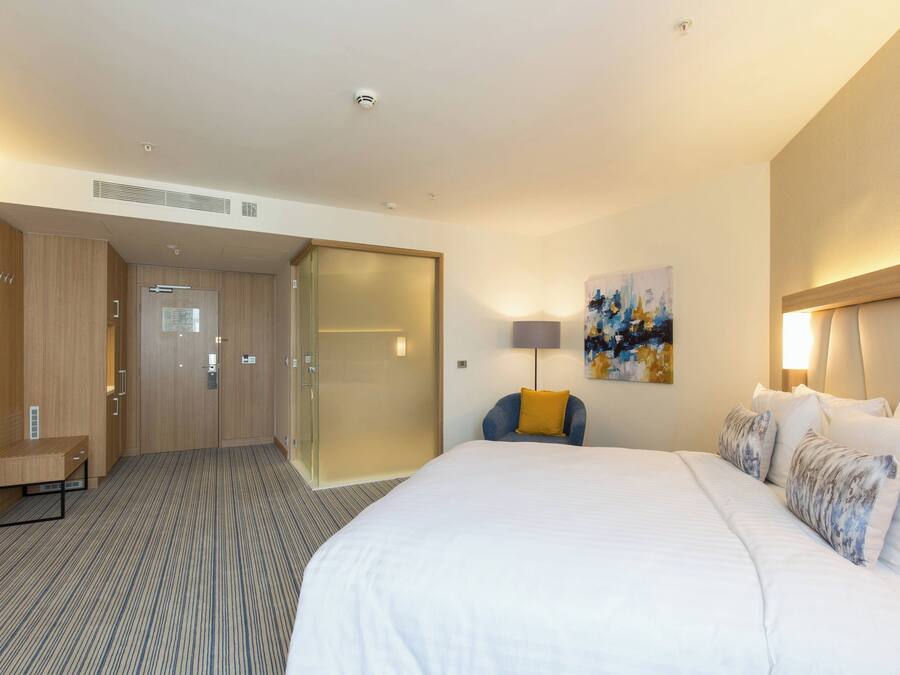 A hotel room with a large bed, a desk, and a chair. There is a painting on the wall and a lamp on the desk.