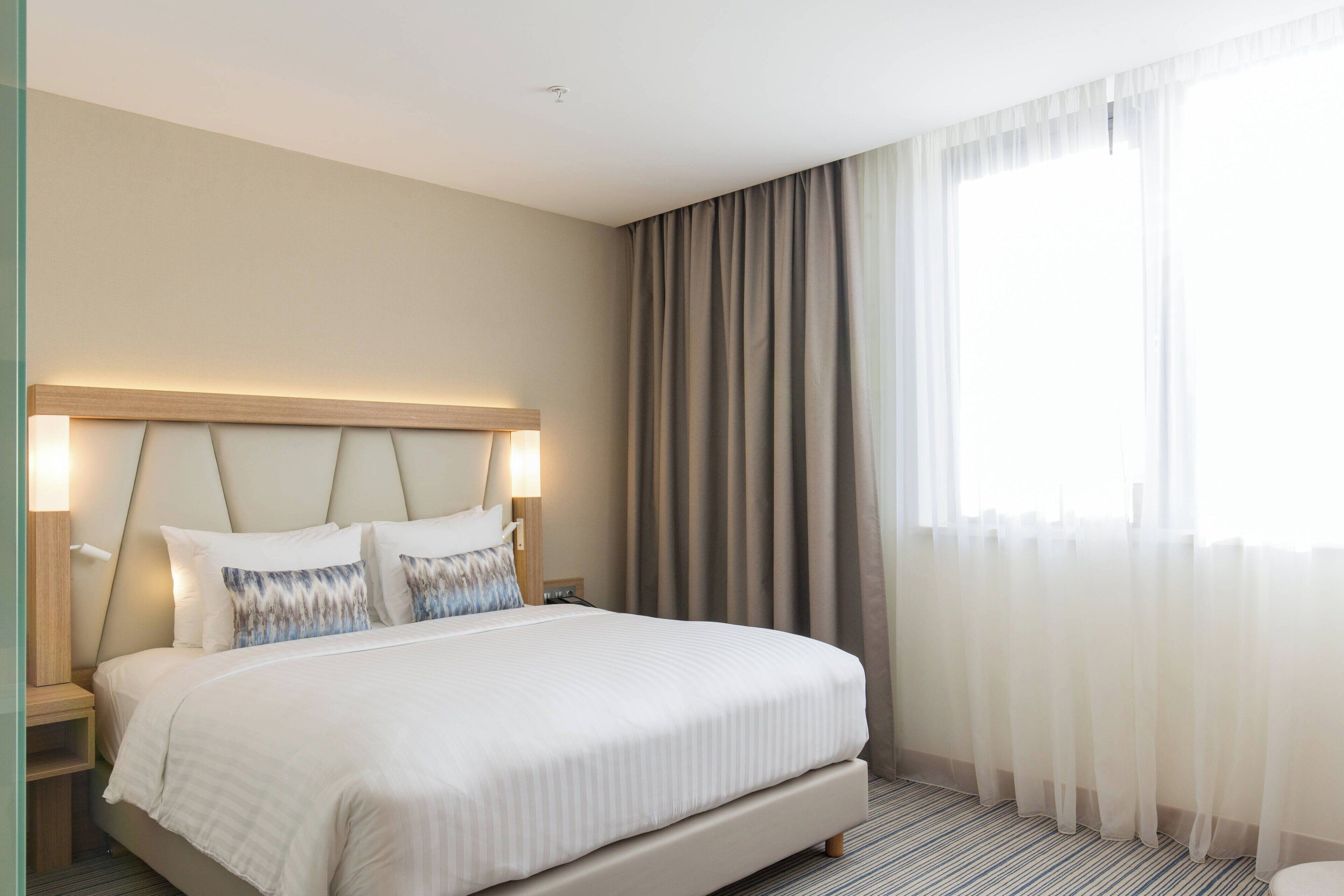 Habitació d'hotel amb un llit gran, un capçal integrat amb il·luminació, una taula de nit i una finestra amb cortines translúcides.