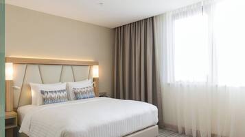 Habitació d'hotel amb un llit gran, un capçal integrat amb il·luminació, una taula de nit i una finestra amb cortines translúcides.