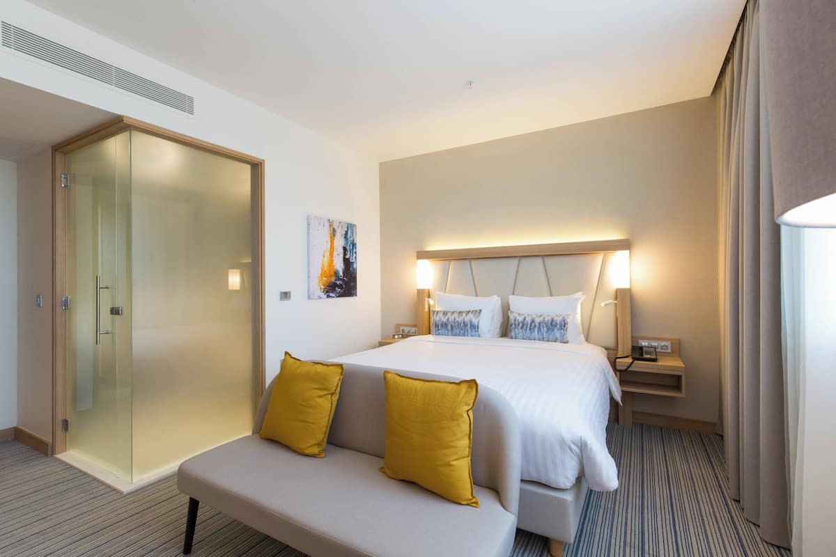 A hotel room with a large bed, a sofa, a glass-enclosed shower, and a painting on the wall.