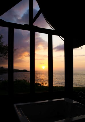A room with a large window overlooking a sunset over the ocean, a bathtub, and a thatched roof structure. - Villa Campuhan (Karangasem)