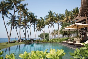 A poolside area with lounge chairs and umbrellas, surrounded by palm trees and a view of the ocean. - Villa Campuhan (Karangasem)