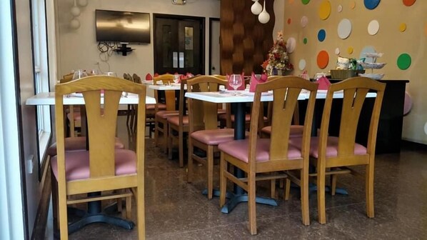 A dining area with wooden chairs and tables, a television mounted on the wall, and a colorful wall decoration.