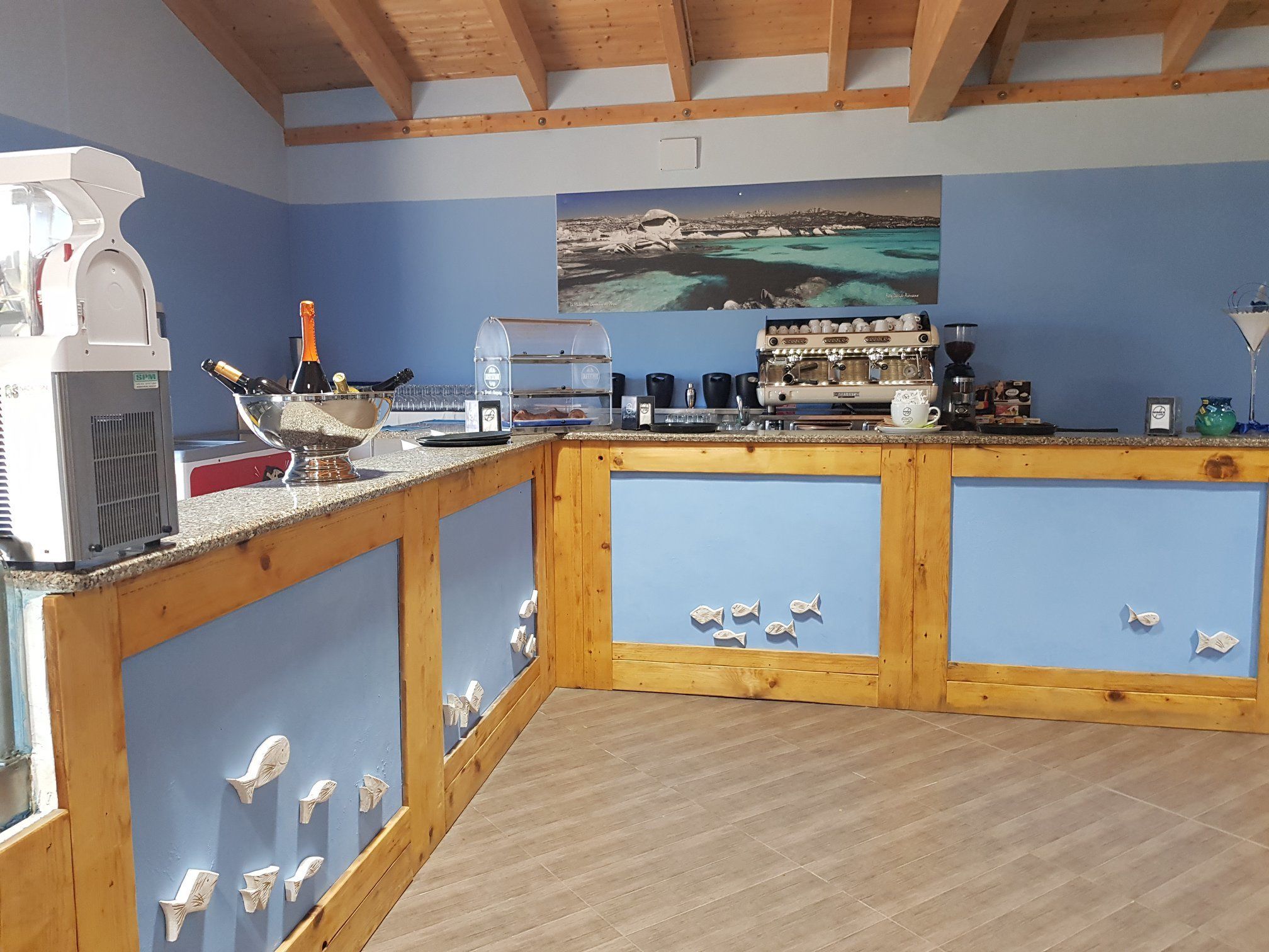 Un espace bar équipé d’une machine à café, d’une vitrine réfrigérée et d’un tableau représentant un paysage côtier.