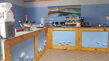 Un espace bar équipé d’une machine à café, d’une vitrine réfrigérée et d’un tableau représentant un paysage côtier.
