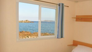 A room with a bed, a window with a view of the sea, and a wooden shelf. - Camping Village Abbatoggia (La Maddalena)