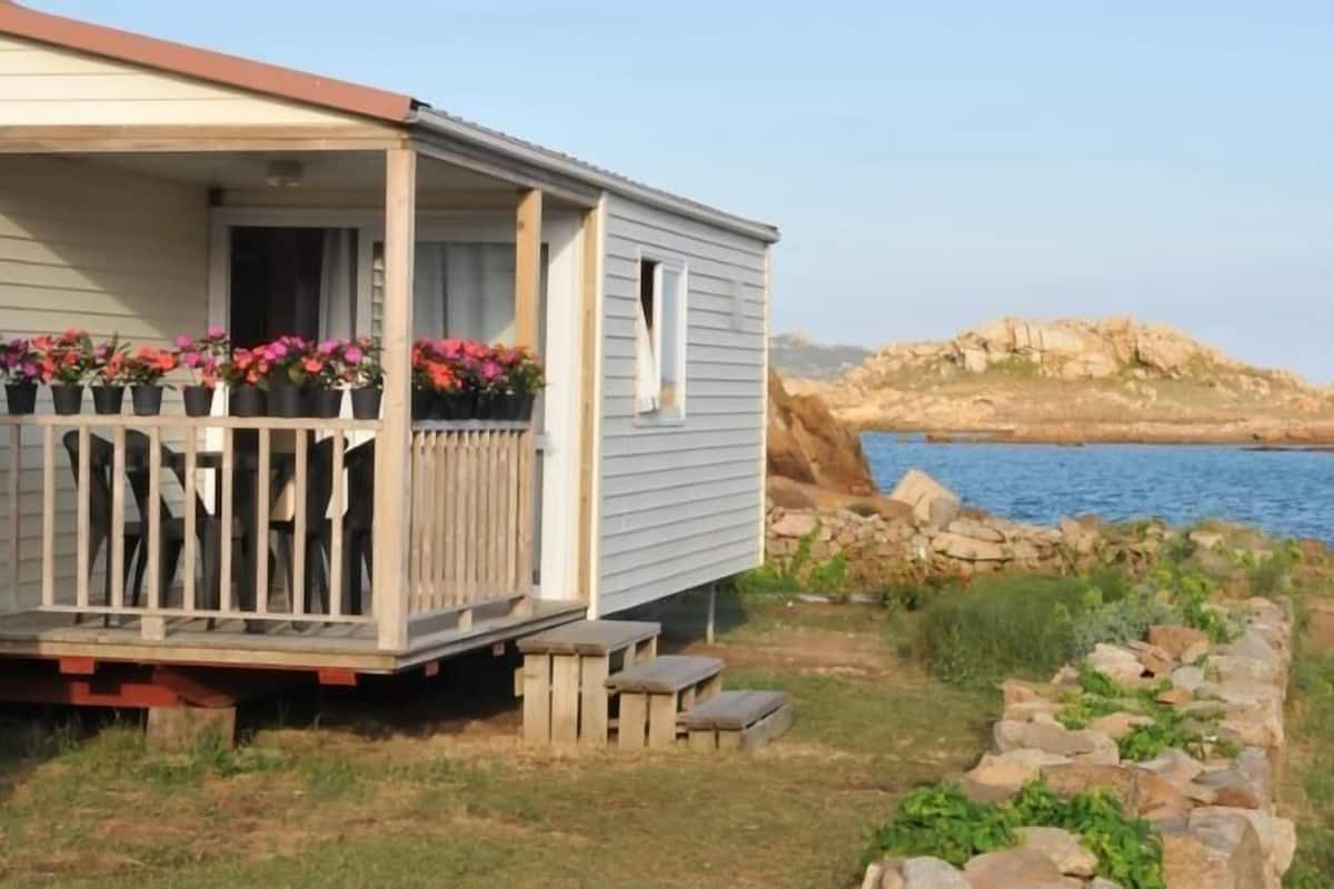 Een gelijkvloerse stacaravan met een veranda, bloempotten en uitzicht op het water en het rotsachtige landschap.