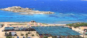 Kystområde med solpaneler, en klippeø og et lille bebyggelse med huse og parkerede køretøjer.