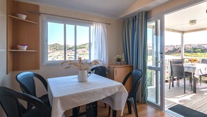 A dining area with a table set for two, a vase with dried plants, and a view of a rocky landscape through large windows. - Camping Village Abbatoggia (La Maddalena)