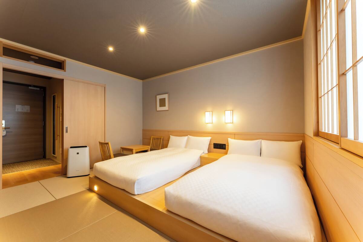 A hotel room with two beds, a bedside table, a chair, a small table, and a wall-mounted light fixture.