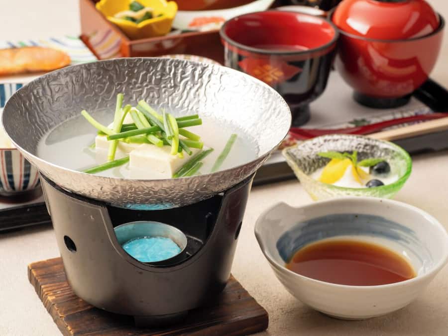 A traditional Japanese hot pot setup with tofu and green onions, accompanied by bowls of soy sauce and pickled vegetables.