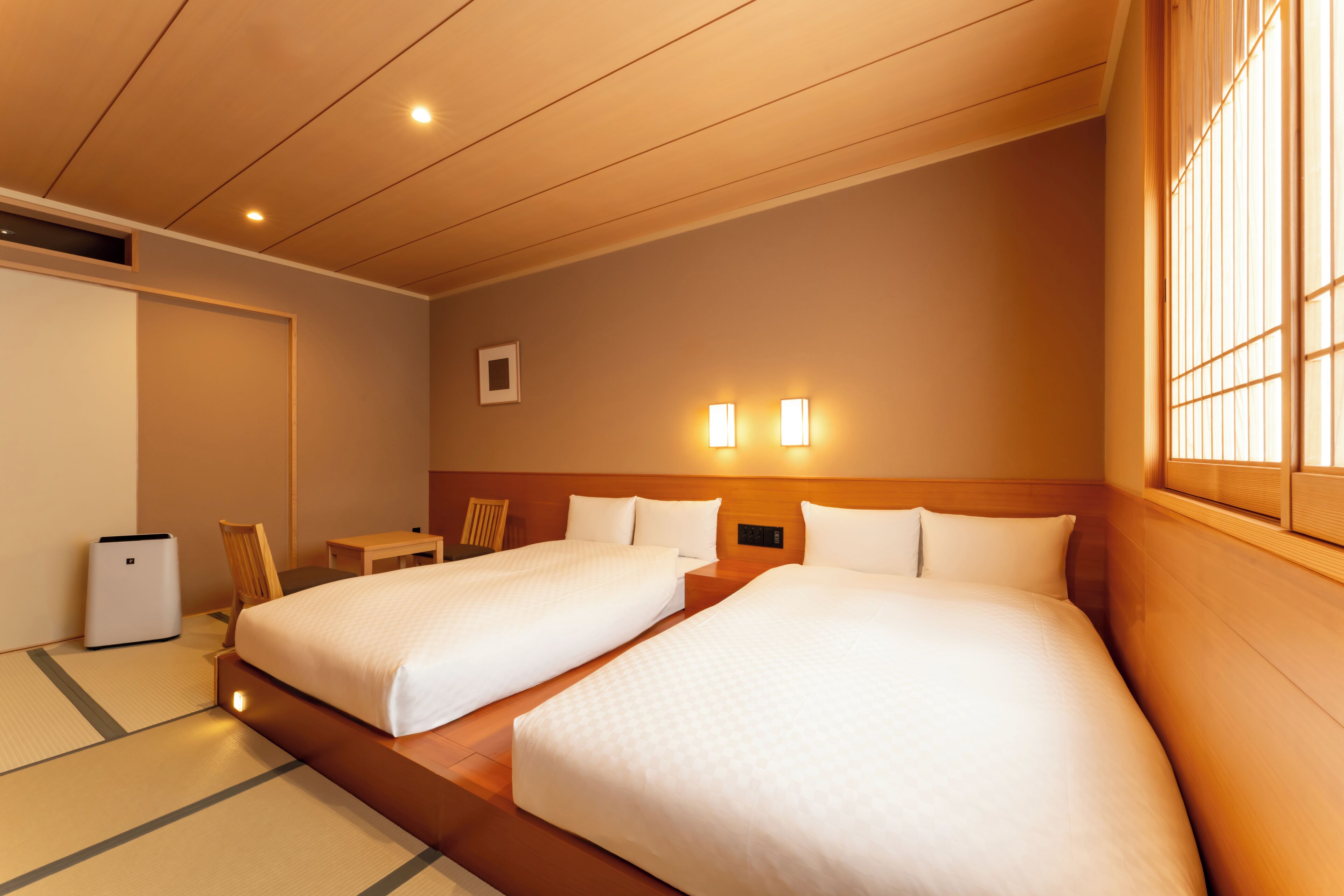 A hotel room with two beds, a small table, a chair, and a bedside table.