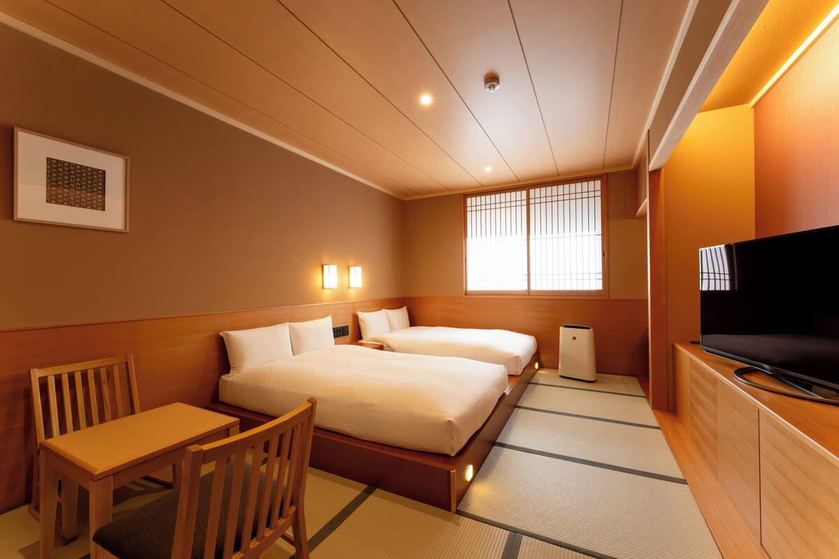 A hotel room with two beds, a desk, a chair, a television, and a window with blinds.
