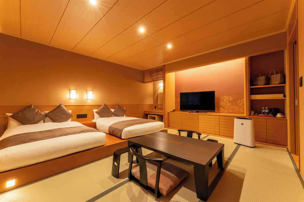 A hotel room with two beds, a dining table, a TV, and a small kitchen area.