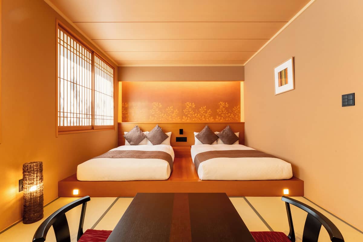 A modern hotel room with two beds, a large window with vertical blinds, a wall-mounted light, and a small picture frame.