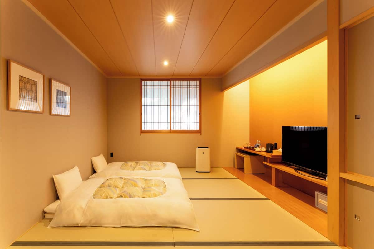 A traditional Japanese room with tatami flooring, a bed with white bedding, a television on a wooden stand, and a sliding door.