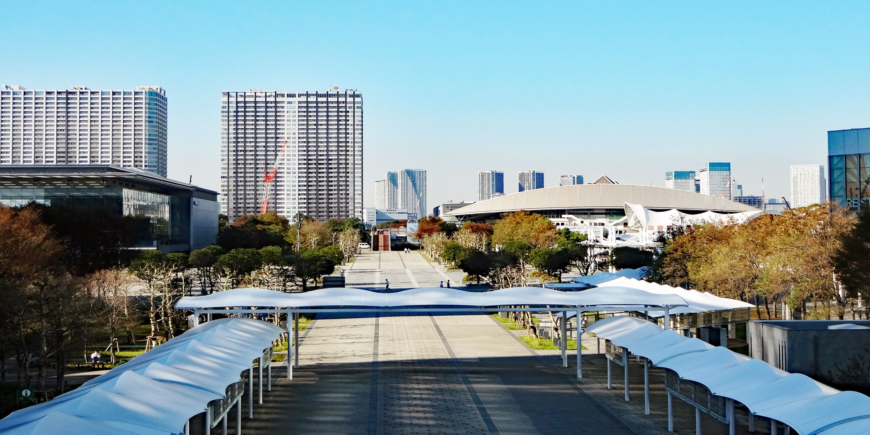 A cityscape with modern buildings, a covered walkway, and a large stadium.