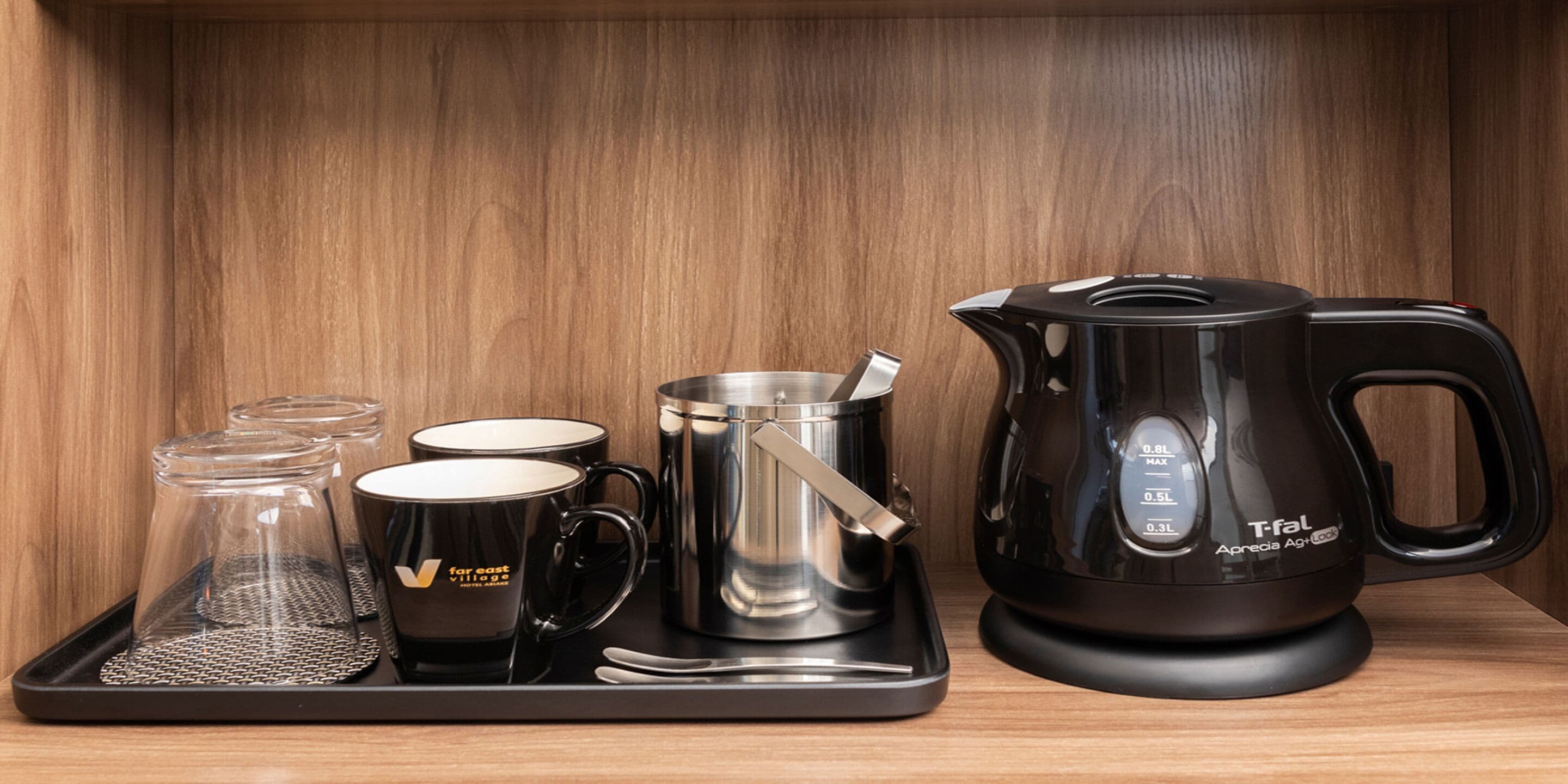 Una estación de café con un hervidor eléctrico Tefal, una cafetera de acero inoxidable, dos tazas negras y una jarra de vidrio sobre una bandeja.