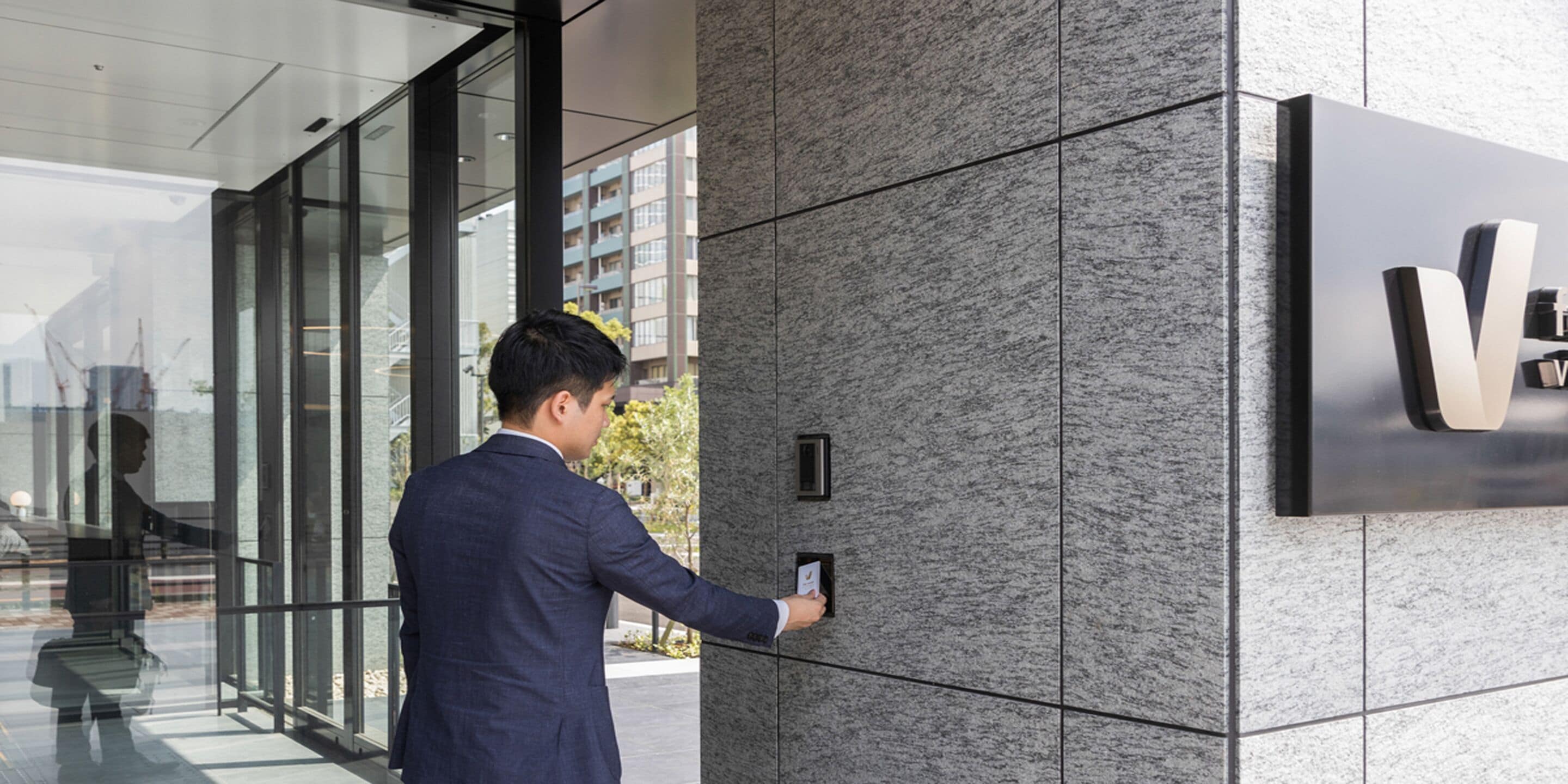A man in a suit interacting with a modern building entrance, featuring a glass facade and a prominent logo on the wall.