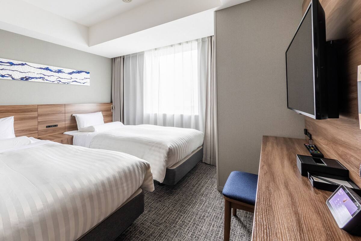 A hotel room with two beds, a desk with a phone, a TV, and a window with curtains.