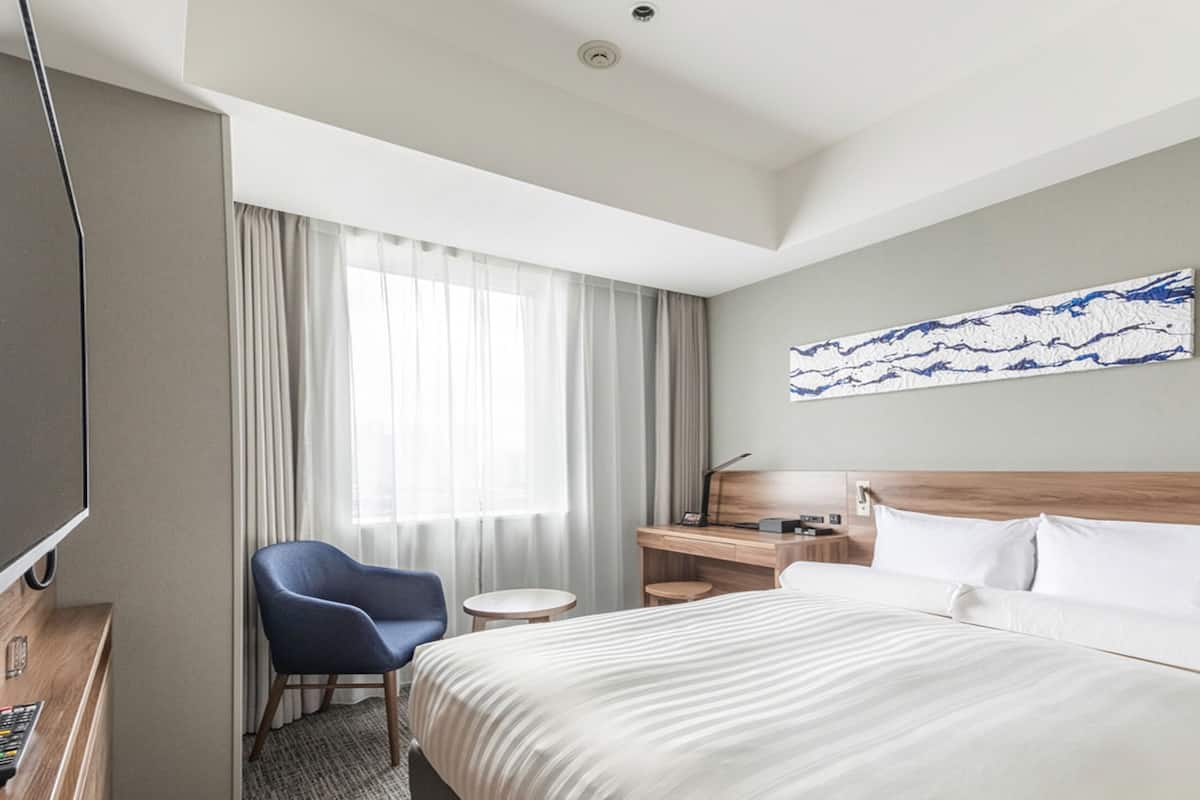A hotel room with a bed, a desk, a chair, a TV, and a window with curtains.