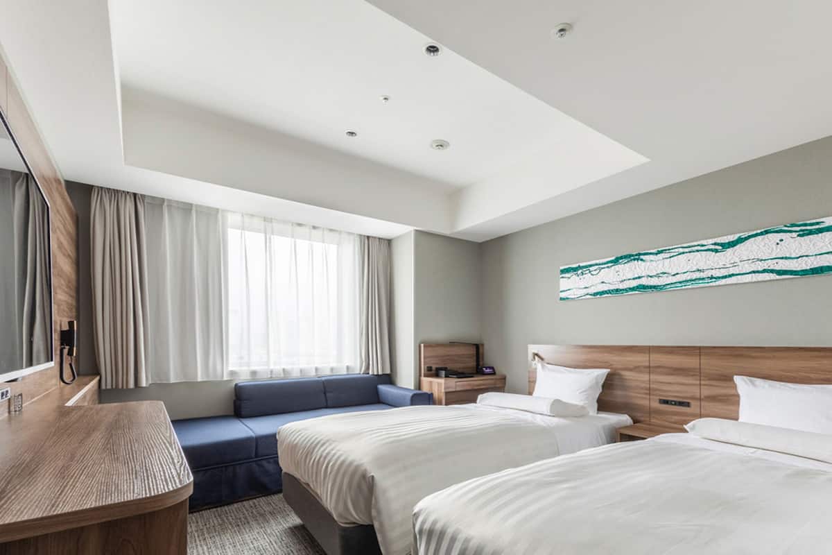 A modern hotel room with two beds, a large window with curtains, a wooden desk, and a wall-mounted artwork.