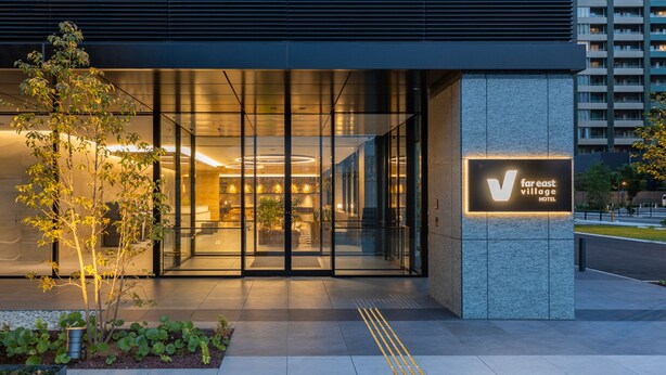 Ingresso moderno di un hotel con vetrate, insegna con la scritta 'V Village Hotel' e area giardino curata.