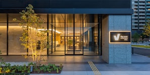 A modern hotel entrance with glass doors, a sign for 'V Village Hotel', and a landscaped garden area. - Far East Village Hotel Ariake, Tokyo (Tokyo)