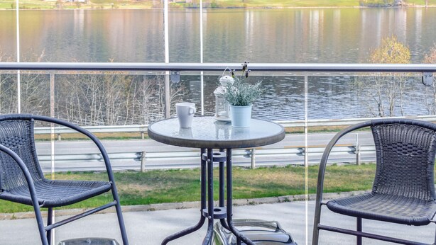 A balcony with two wicker chairs, a small round table, and a potted plant. A view of a lake and distant buildings is visible through the railing. - Stunning apartment in Vossestrand (Vossestrand)