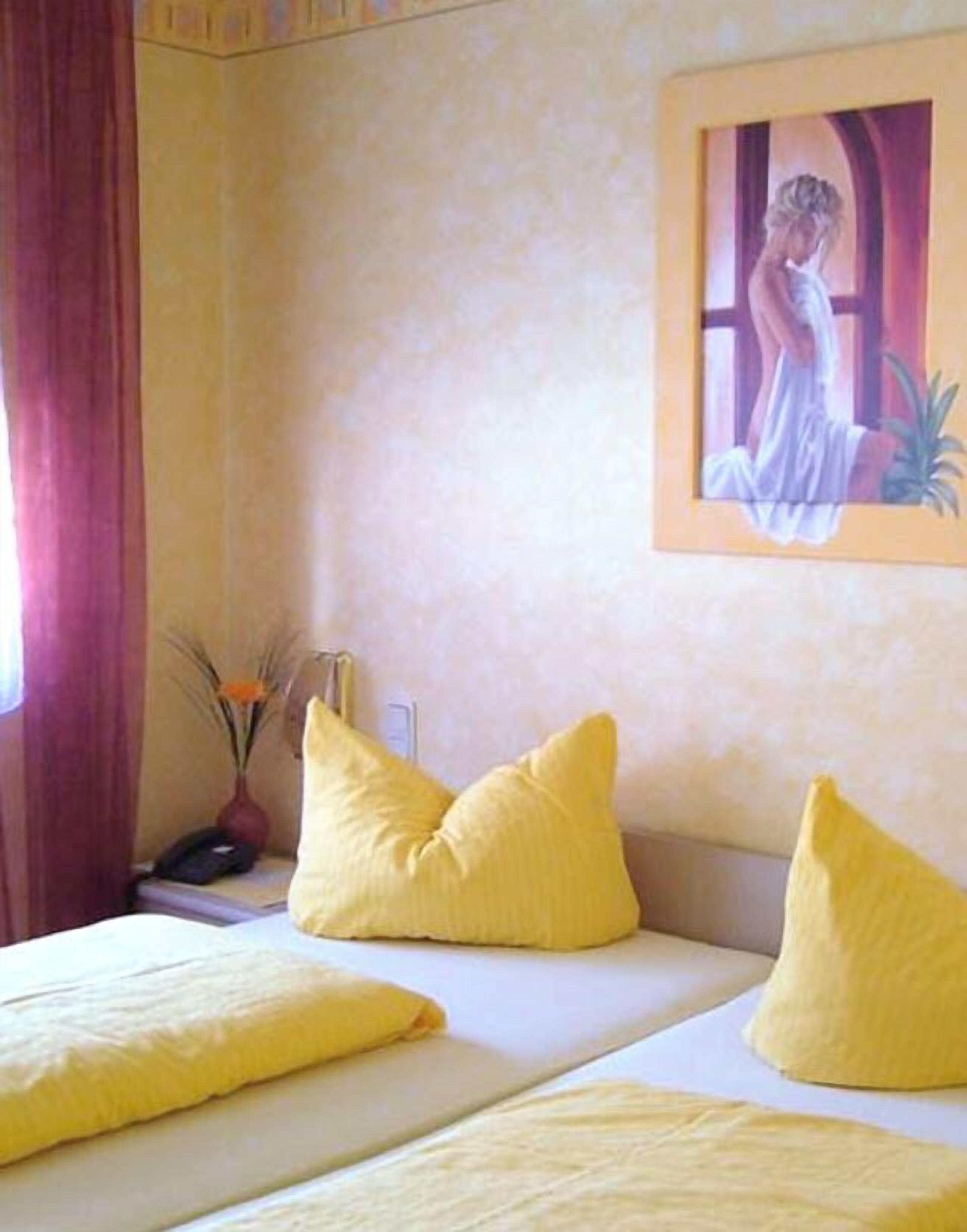 Hotellihuone, jossa on sänky, kaksi keltaista tyynyä, seinällä taulu ja maljakko, jossa on kukkia.