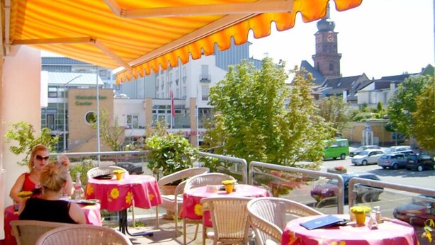 An outdoor seating area with tables and chairs under a yellow awning, overlooking a street with cars and buildings. - Stadt Cafe Pension Grünstadt (Gruenstadt)