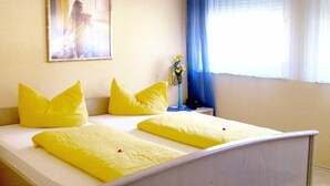 A bed with yellow bedding and pillows, a bedside table with a vase of flowers, and a window with blue curtains. - Stadt Cafe Pension Grünstadt (Gruenstadt)