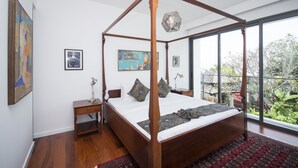 Ein Bett mit Baldachin und Holzrahmen, ein Nachttisch mit Lampe und Blick auf Grün hinter großen Fenstern.