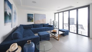 Ein modernes Wohnzimmer mit einem blauen Ecksofa, einem Glastisch und einem großen Fenster mit Blick auf Gartenmöbel.