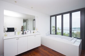 Een moderne badkamer met een grote spiegel, twee wasbakken, een bad en uitzicht op zee door grote ramen.