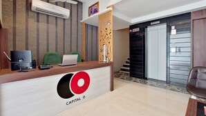 A reception desk with a logo, a computer, and a green chair. There is a wall-mounted air conditioner and a decorative panel. - Capital O 30351 S B Excellency (Jodhpur)