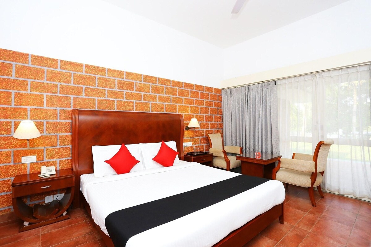 A hotel room with a brick-patterned wall, a wooden bed, a bedside table with a lamp, a chair, and a window with curtains.