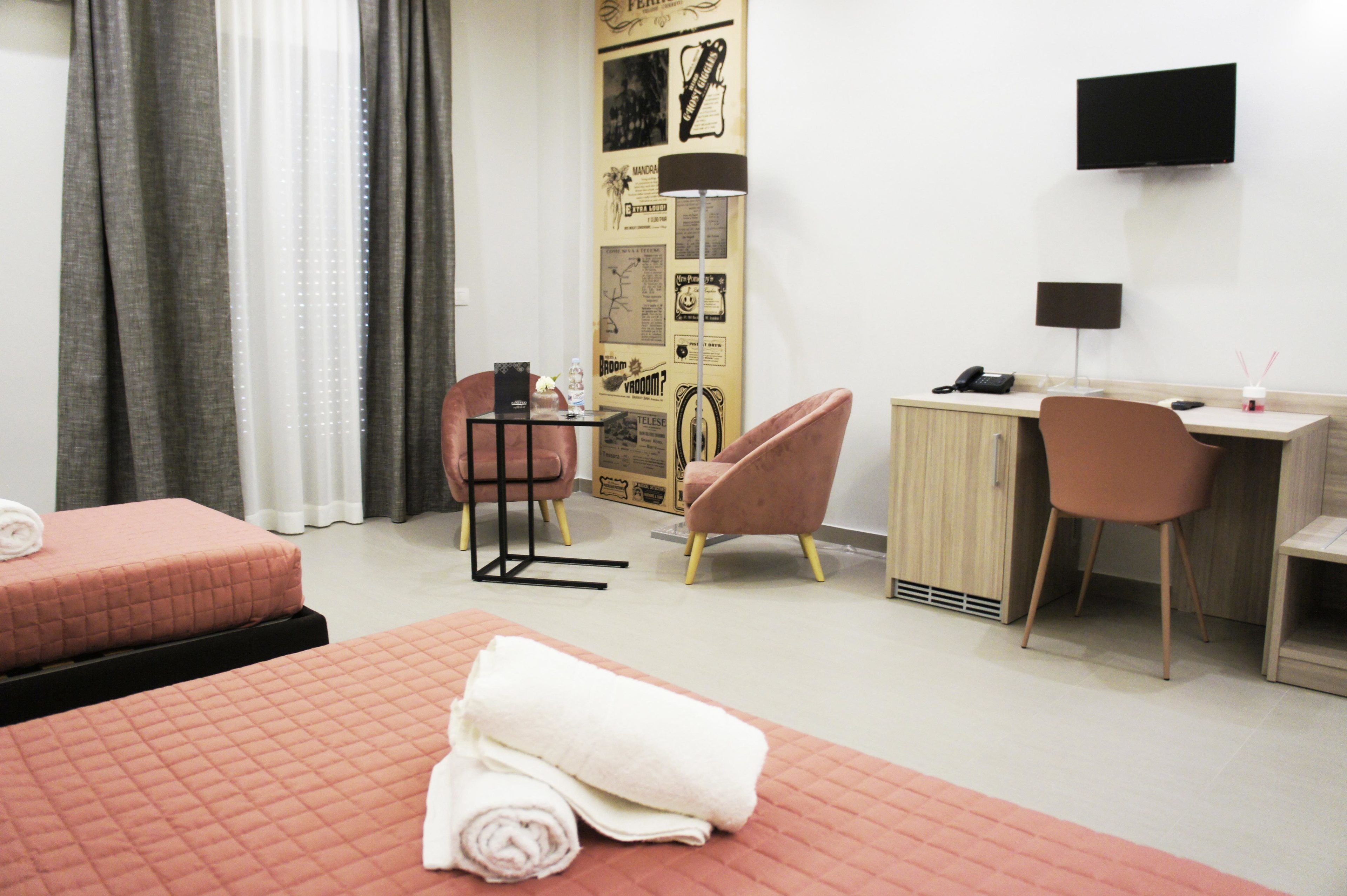 A hotel room with two beds, a desk, a chair, a TV, and a wall with posters.