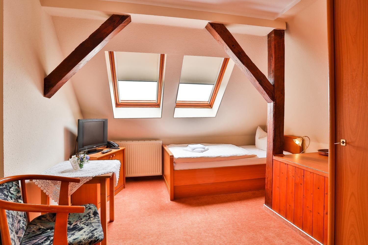 A small, cozy room with a bed, a desk, a chair, and a television. There are two windows providing natural light.