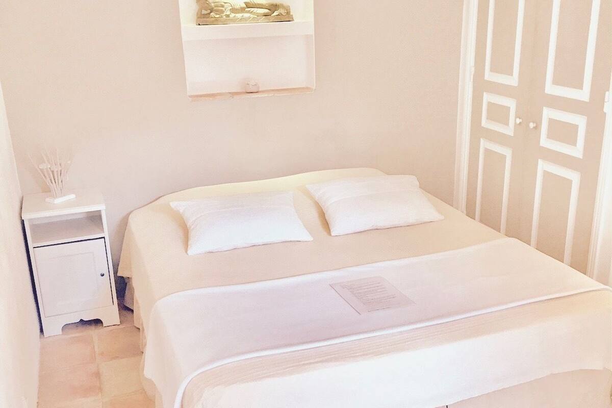 Una cama bien hecha con ropa blanca en una habitación con suelo de baldosas y una mesita de noche blanca con una lámpara.