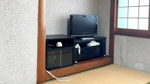A room with a television on a wooden stand, a small cabinet below it, and a window with a grid pattern. - Business Ryokan Wakakusa (Tsu)