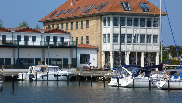 A marina with several boats docked, a building with a red roof, and a clear blue sky. - Wohnung Residenz am Wasser (Karlshagen)