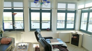 A spacious living room with a view of a body of water through large windows, a black leather sofa, a small table, and a radiator. - Wohnung Residenz am Wasser (Karlshagen)