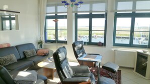 A living room with a sofa, two chairs, a coffee table, and a view of a marina through large windows. - Wohnung Residenz am Wasser (Karlshagen)