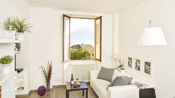 A modern living room with a white sofa, a small table with a bottle and glasses, a potted plant, and a window with a view of greenery. - Breathtaking view from a charming apartament (Menaggio)